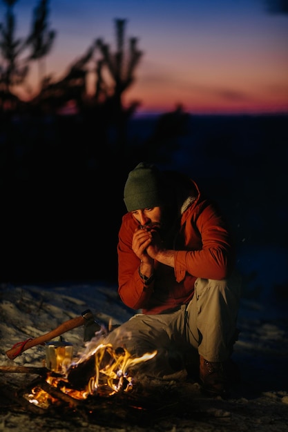 Un homme est assis près d'un feu de camp se réchauffant près du feu dans une forêt d'hiver au coucher du soleil