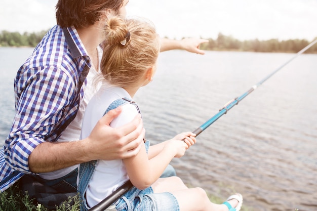 L'homme est assis avec sa fille au bord de la rivière et pointe vers l'avant. Fille tient une canne à pêche et pêche.