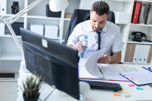 Un homme est assis à une table dans le bureau, travaillant avec des documents et tenant une bouteille d’eau à la main.