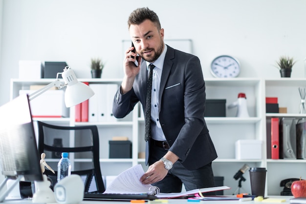 Un homme est debout près d'une table dans le bureau, parlant au téléphone et feuilletant des documents.