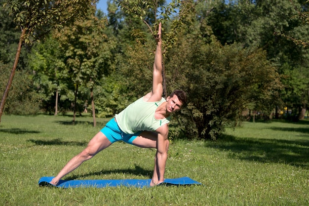 L'homme étire les mains et les jambes sur un tapis bleu dans le parc. Sportif avec corps musclé sur l'herbe verte. Athlète faisant de l'exercice le jour d'été ensoleillé en plein air. Sport, yoga, pilates, fitness. Concept de mode de vie sain