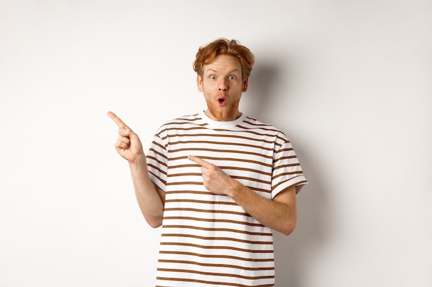 Homme étonné aux cheveux roux vérifiant l'offre promotionnelle, pointant vers la gauche le logo et souriant à la caméra, debout sur fond blanc.