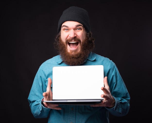 Homme étonné gai avec barbe montrant un écran vide blanc sur ordinateur portable.