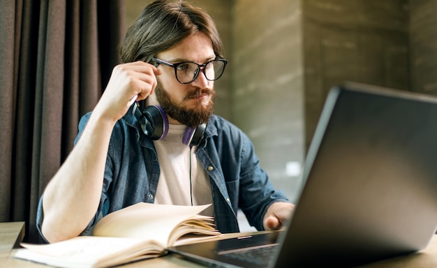 Homme étudiant barbu dans des écouteurs portant des lunettes à l'aide d'un ordinateur portable le soir à l'appartement