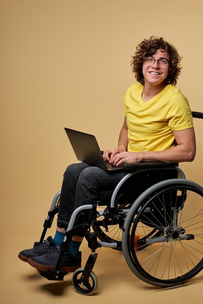 homme étudiant handicapé prépare pour examen à l'aide d'un ordinateur portable, assis sur un fauteuil roulant isolé en studio. portrait. éducation en ligne pour les personnes handicapées