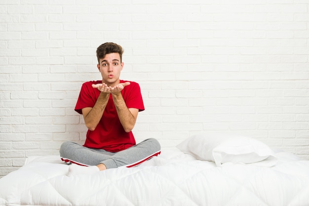 Homme étudiant jeune adolescent sur le lit pliant les lèvres et tenant les paumes pour envoyer un baiser aérien.