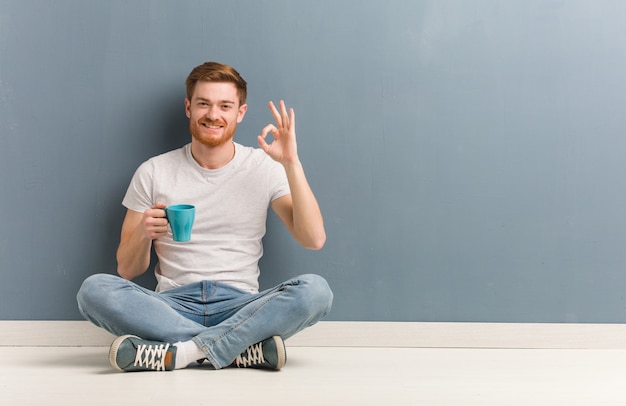 Homme étudiant jeune rousse assis sur le sol gai et confiant faisant geste ok. Il tient une tasse de café.