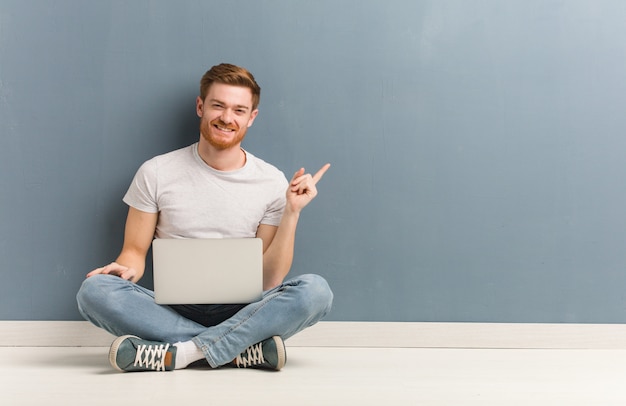 Homme étudiant jeune rousse assis sur le sol pointant sur le côté avec le doigt. Il tient un ordinateur portable.
