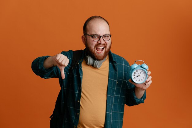 Homme étudiant en tenue décontractée portant des lunettes avec un casque tenant un réveil regardant la caméra avec une expression agacée montrant le pouce vers le bas debout sur fond orange