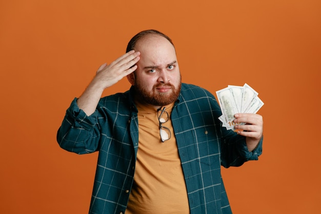 Homme étudiant en tenue décontractée tenant de l'argent en regardant la caméra confondue avec la main sur la tête pour erreur debout sur fond orange