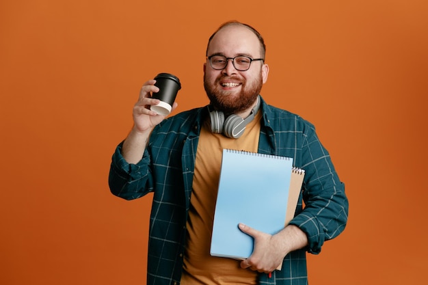 Homme étudiant en vêtements décontractés portant des lunettes avec des écouteurs tenant des cahiers et une tasse de café heureux et positif souriant joyeusement debout sur fond orange