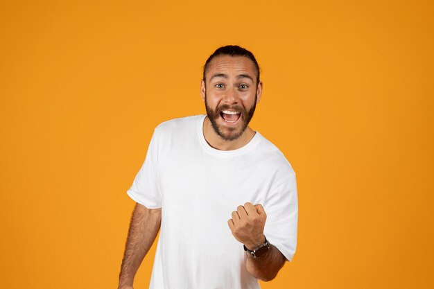 Un homme européen adulte souriant avec une barbe en t-shirt blanc se réjouit de la bonne nouvelle, la victoire célèbre le succès