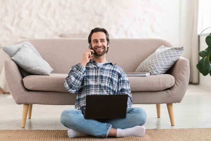 Photo un homme européen souriant du millénaire en vêtements domestiques avec du chaume est assis sur des appels au sol par téléphone fonctionne sur pc