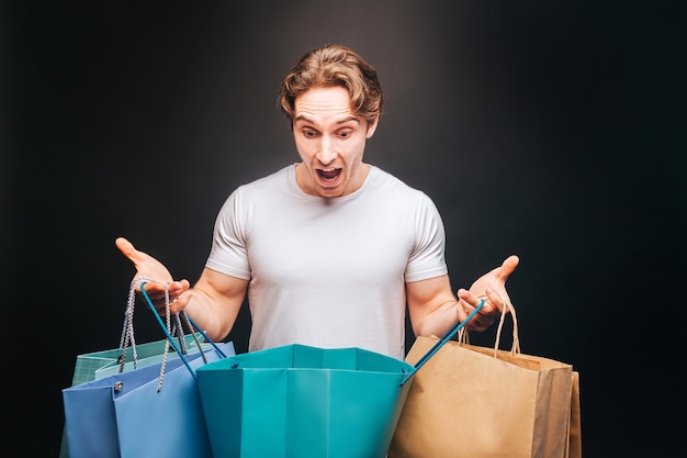 Homme européen surpris regardant dans un sac à provisions ouvert. Jeune beau mec porte des vêtements décontractés avec des sacs de magasin. Concept de shopping. Isolé sur fond sombre. Tournage en studio. Espace de copie