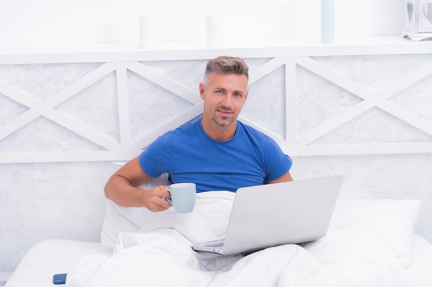 Un homme éveillé boit du café à l'aide d'un ordinateur portable au lit le matin