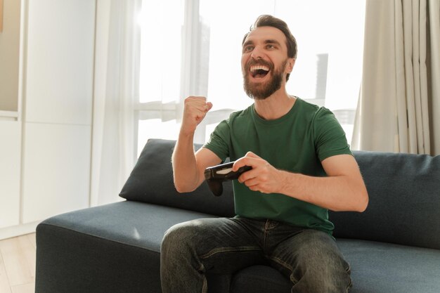 Homme excité assis sur un canapé confortable dans un appartement jouant à des jeux vidéo faisant le geste du gagnant