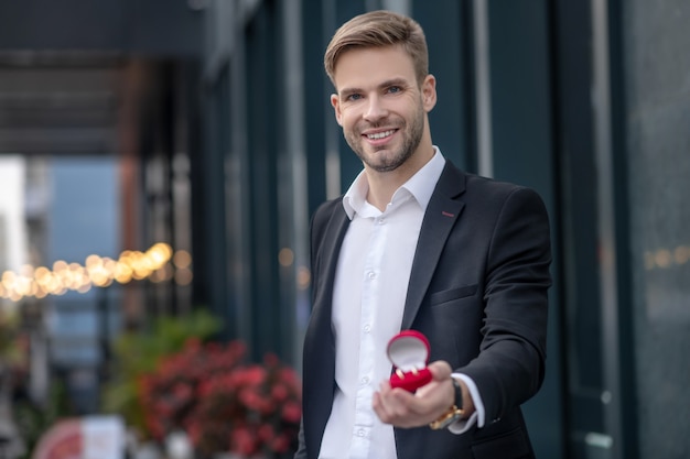 Homme excité en costume noir montrant une boîte avec bague de fiançailles