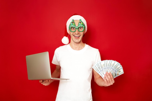 Homme excité dans un chapeau et des lunettes de nouvel an, des arbres de Noël tient un ordinateur portable et un ventilateur d'argent dans ses mains