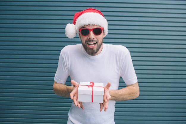 Un homme excité et étonnant se tient debout et attend avec impatience. Il porte un chapeau et des lunettes de Noël. Aussi le gars tient le présent dans les mains. Isolé sur rayé