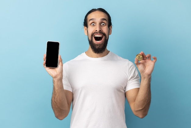 Homme excité étonné avec barbe portant un t-shirt blanc tenant un téléphone portable et du bitcoin doré