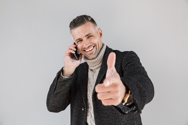 Homme excité séduisant portant un manteau debout isolé sur un mur gris, parlant au téléphone portable, pointant le doigt vers l'avant