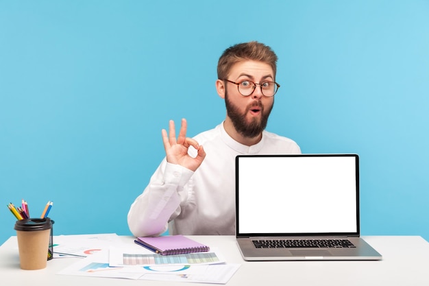 Homme excité surpris avec barbe en chemise blanche et lunettes montrant un geste correct avec les doigts démontrant un ordinateur portable à écran blanc assis sur le lieu de travail Studio intérieur tourné isolé sur fond bleu