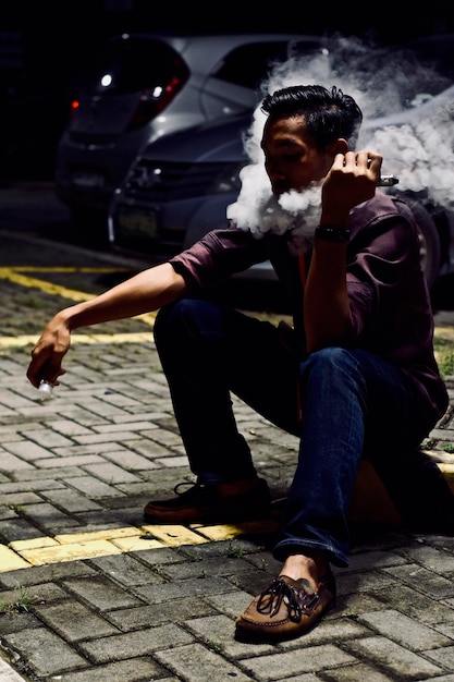 Photo un homme exhale de la fumée alors qu'il est assis sur le trottoir la nuit.