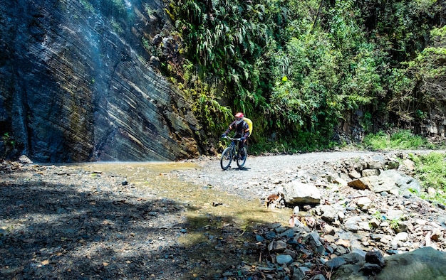 Homme faisant du vélo sur la route de la mort bolivienne ensoleillée à proximité d'un énorme rocher avec cascade