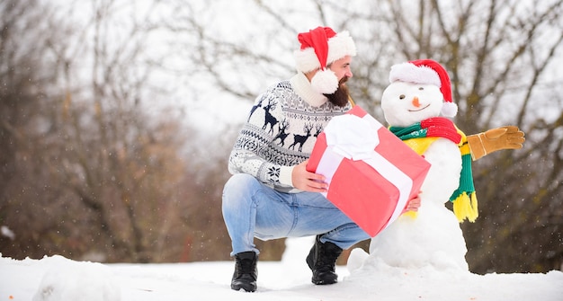 L'homme a fait un bonhomme de neige. Bonnet de Noel homme s'amusant à l'extérieur. Guy visage heureux nature enneigée fond. Hipster avec barbe tenir la boîte-cadeau. Notion de surprise. Jeu d'hiver. Activité hivernale. Vacances d'hiver.