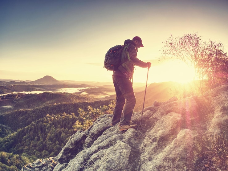 Photo homme sur une falaise rocheuse grimpant avec des bâtons de trekking et un coucher de soleil à dos derrière l'homme