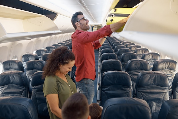 Photo un homme de famille joyeux met ses bagages à main dans le compartiment tout en voyageant avec sa femme et sa petite fille en avion. vacances en famille, concept de transport