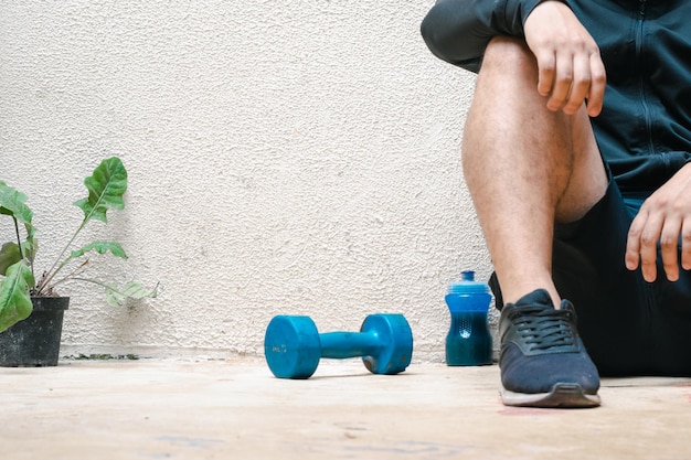 Homme fatigué assis sur le sol à côté de l'haltère bleu et d'une bouteille d'eau après l'exercice