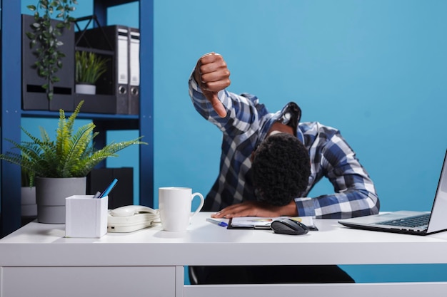 Homme fatigué endormi montrant un geste d'aversion tout en posant la tête sur le bureau. Un employé de l'entreprise qui travaille dur, affligé et épuisé, s'endort sur le bureau tout en montrant le symbole de désapprobation avec la main.