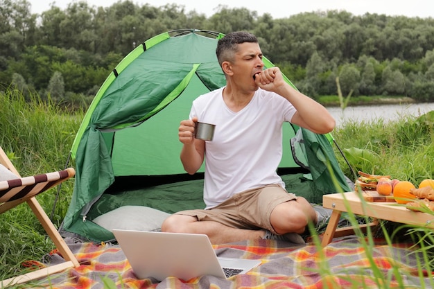 Un homme fatigué travaillant sur un ordinateur portable à l'extérieur un pigiste a la capacité de travailler de n'importe où dans le monde assis près de la tente et de boire du café a besoin d'énergie se sent somnolent bâillant couvrant la bouche avec la main