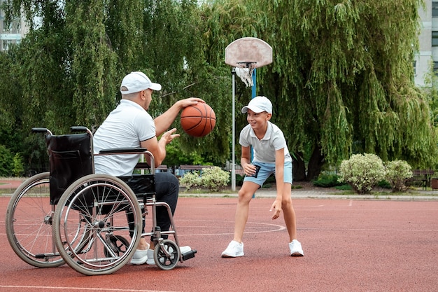 Un homme en fauteuil roulant joue au basket avec son fils sur le terrain de sport. Parent handicapé, enfance heureuse, concept de personne handicapée.