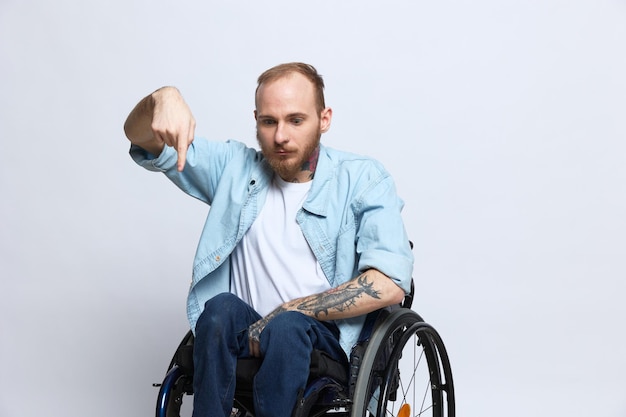 Un homme en fauteuil roulant et pointant un doigt vers l'espace de copie avec des tatouages sur ses mains est assis sur un fond de studio gris concept de santé une personne handicapée une personne réelle