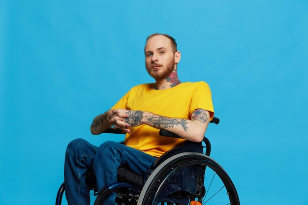 Un homme en fauteuil roulant a des problèmes avec le système musculo-squelettique regarde la caméra dans un t-shirt avec des tatouages sur ses bras est assis sur un fond de studio bleu concept de santé de personne réelle pleine vie