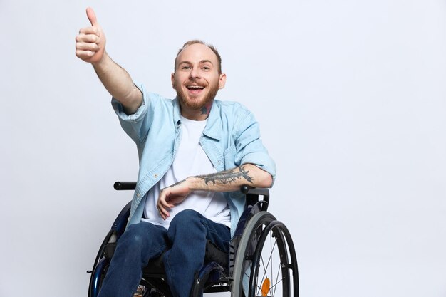 Un homme en fauteuil roulant regarde la caméra montre un pouce levé le bonheur avec des tatouages sur ses mains est assis sur un fond de studio gris concept de santé homme handicapé