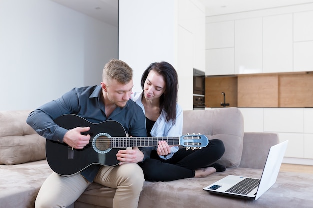 Homme et femme apprenant à jouer de la guitare ensemble à l'aide d'un ordinateur portable, jeune couple ayant un bon moment ensemble à la maison assis sur un canapé