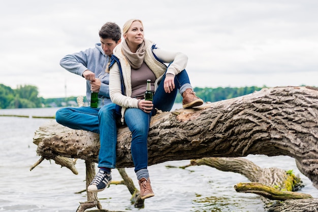 Homme et femme assis sur le tronc près au bord du lac et boire de la bière en bouteille