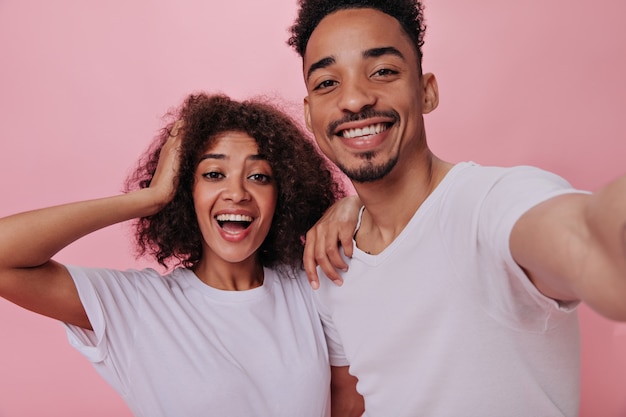 Un homme et une femme aux yeux bruns en T-shirts blancs prennent un selfie sur un mur rose