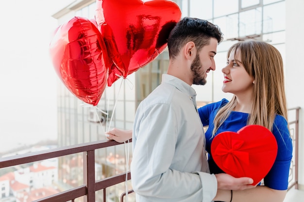 Homme et femme avec cadeau et ballons en forme de coeur rouge sur balcon à la maison