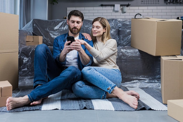 Un homme et une femme dans un nouvel appartement un couple marié assis sur le sol près de boîtes en carton utilisant le téléphone pour faire des achats en ligne en choisissant des meubles