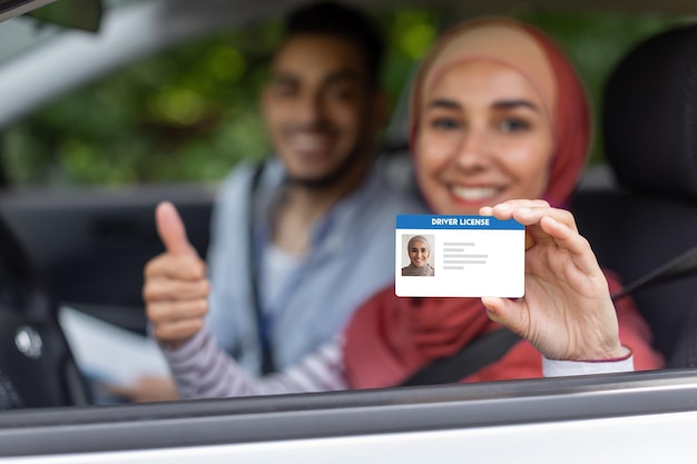Photo un homme et une femme heureux du moyen-orient en hijab montrent un permis de conduire et le pouce vers le haut dans la fenêtre ouverte de la voiture