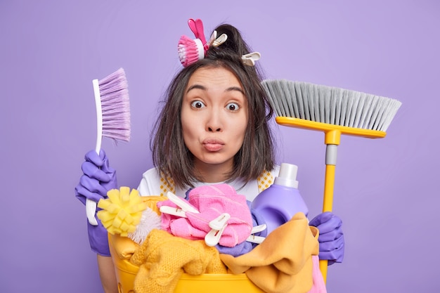 homme femme de ménage tient une brosse et un balai utilise des agents de nettoyage des blanchisserie à la maison porte des gants de protection en caoutchouc se tient près d'un panier plein d'articles à laver isolé sur violet