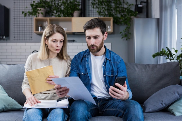 Un homme et une femme réfléchis paient des factures à la maison utilisent l'application au téléphone sérieux au sujet de la paperasse