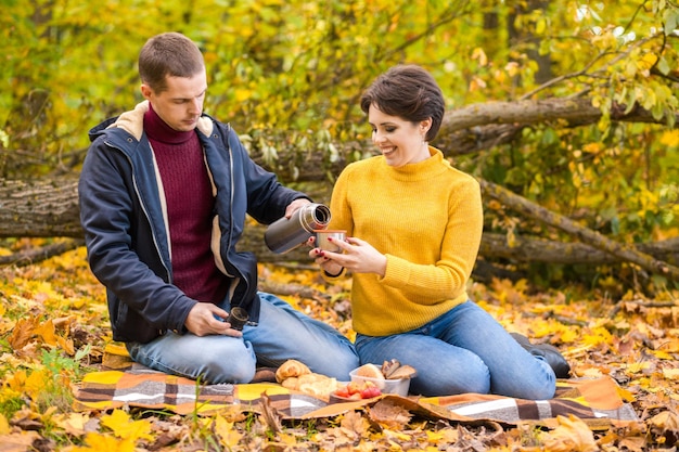 Un homme et une femme s'assoient sur un plaid dans un parc d'automne et boivent une boisson chaude dans un thermos