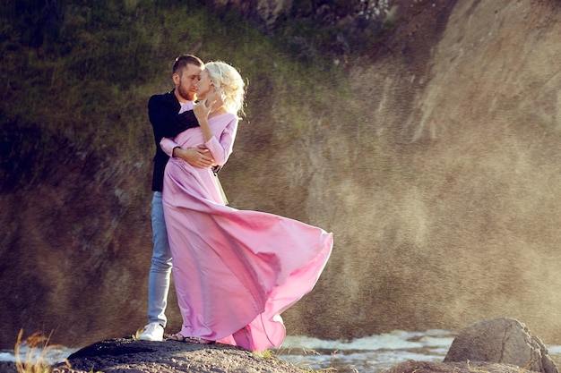 Homme et femme s'embrassant en été au coucher du soleil dans de beaux vêtements. Un couple amoureux se tient sur le rivage sur les rochers au soleil, s'embrassant et s'embrassant dans les éclaboussures d'eau. Robe rose