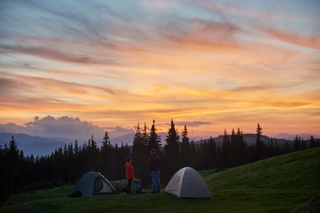 Homme et femme se reposant dans le camping près de deux tentes dans les montagnes lors d'une randonnée avec leurs sacs à dos en profitant du magnifique coucher de soleil