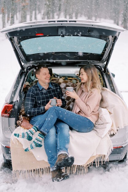 Un homme et une femme souriants assis avec des tasses dans le coffre de la voiture sur des couvertures dans un parc enneigé